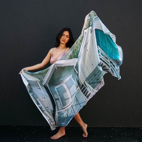 A girls standing up against a dark wall a holding out a cotton linen scarf featuring Portsea Beach boatsheds in greens and greys.