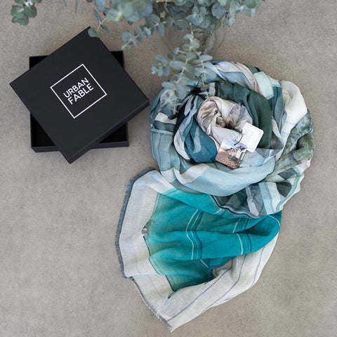 A black gift box and a scrolled up cotton linen scarf featuring Portsea Beach boatsheds in greens and greys.