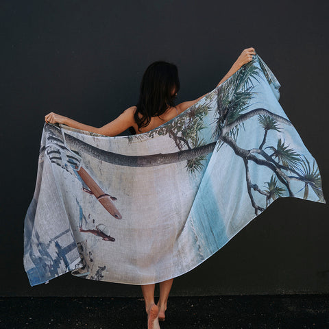 A girl standing up against a black wall holding out cotton linen scarf featuring a design of Little Cove in Noosa.